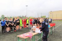 Z prawej strony z przodu policjanta przygotowuje nagrody na stołach. Z lewej strony w tyle stoją uczestnicy turnieju piłkarskiego.
